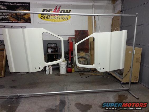 Two white vehicle doors hanging from a metal rack in a workshop.