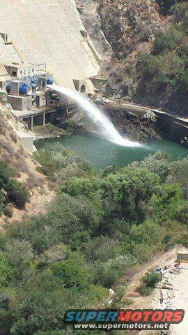 Dam structure with water being released into a reservoir below. Trees and hills surround the area