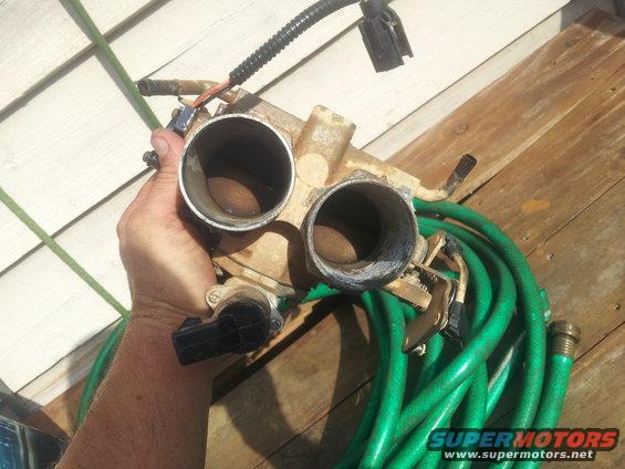 Hand holding a dirty dual throttle body assembly with a green garden hose coiled nearby
