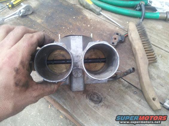 Hand holding a dual throttle body with a wire brush and tools on a wooden surface