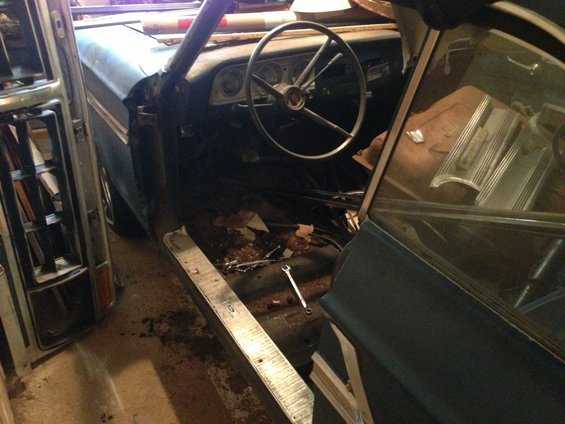 Interior of a car with a steering wheel, dashboard, and open door. Floor is dirty with debris and a wrench