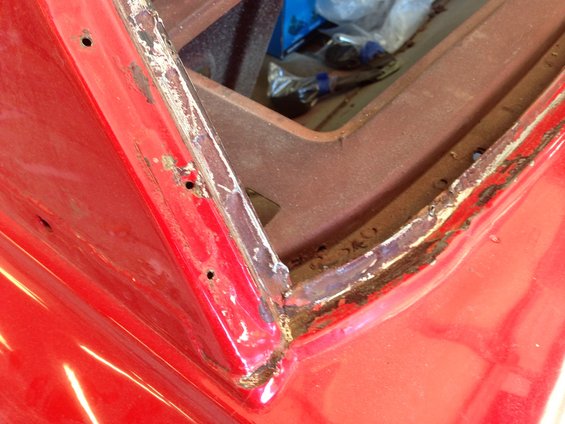 Close-up of red car body panel with rust and holes around the window frame, showing damage and repair needs