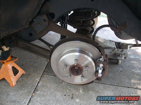 Rear brake rotor and caliper assembly on a vehicle supported by an orange jack stand and a floor jack