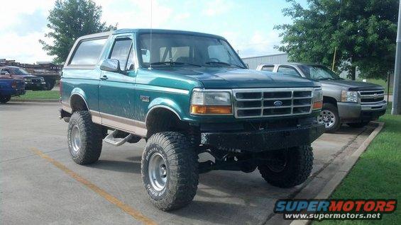 Green 1990s Ford Bronco with lifted suspension and aftermarket bumper parked next to a silver pickup truck