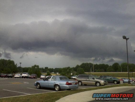 Light blue sedan parked in a lot under a cloudy sky. Other cars are visible in the background