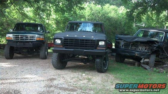 Three Ford trucks parked outside. Center one is black, lifted, with a 1979 grille. Green one on left is newer. Black one on right is wrecked