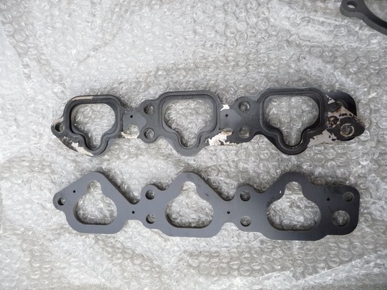 Two engine gaskets on bubble wrap. One gasket is black with white peeling paint and a black rubber seal. The other is dark gray