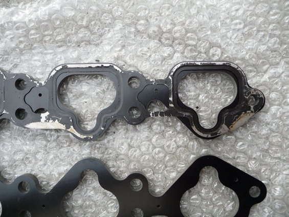 Two engine gaskets, one grey and worn, the other black and new, on a bubble-wrap surface