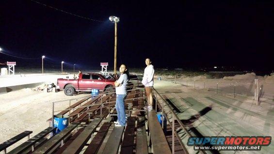 Red pickup truck parked on a dirt track at night, with two people standing on bleachers in the foreground