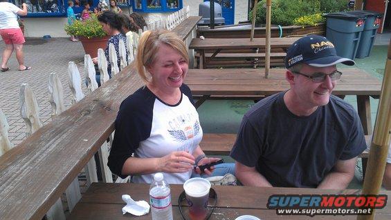 Woman laughs while looking down at her phone, man in a baseball cap sits next to her at a picnic table