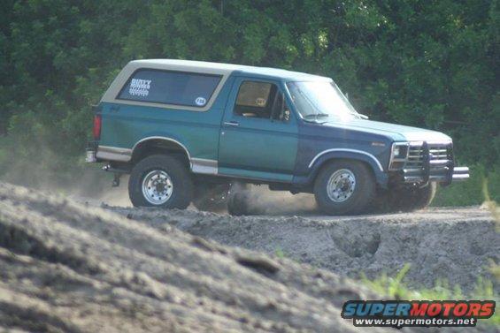 Blue and teal Ford Bronco driving on a dirt road, kicking up dust. Has a tan camper shell