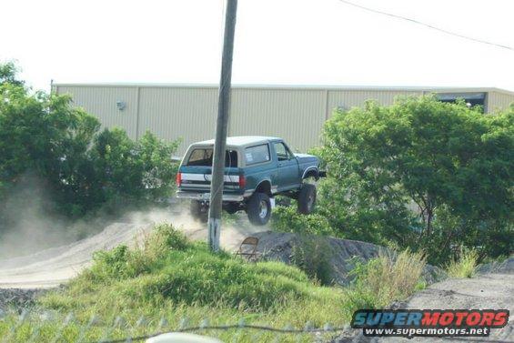 Teal Ford Bronco mid-air jump, rear-wheel drive, lifted suspension, off-road tires, dirt track