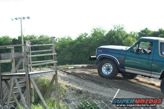 Teal Ford Bronco with tan trim driving on a dirt track near a wooden structure