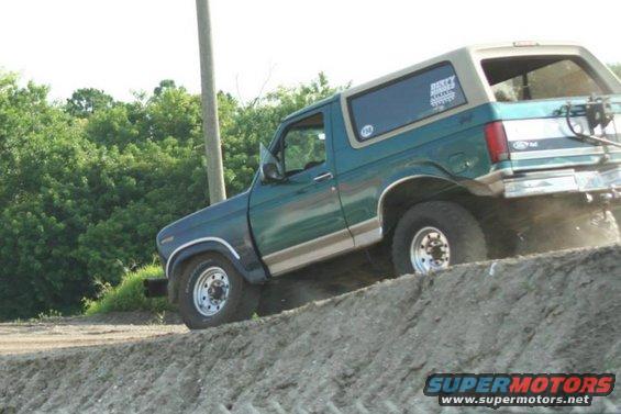Green and tan Ford Bronco driving up a dirt mound, rear tire kicking up dust