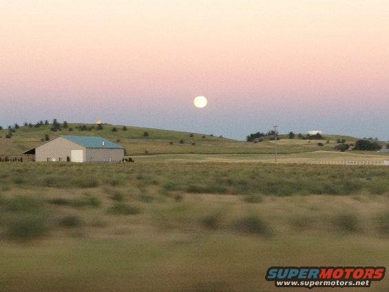 Moon hangs in a pink sky over a barn and rolling hills
