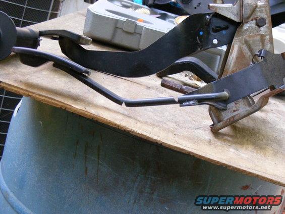 Vehicle pedal assembly with bent metal rod and black plastic components, resting on a wooden surface