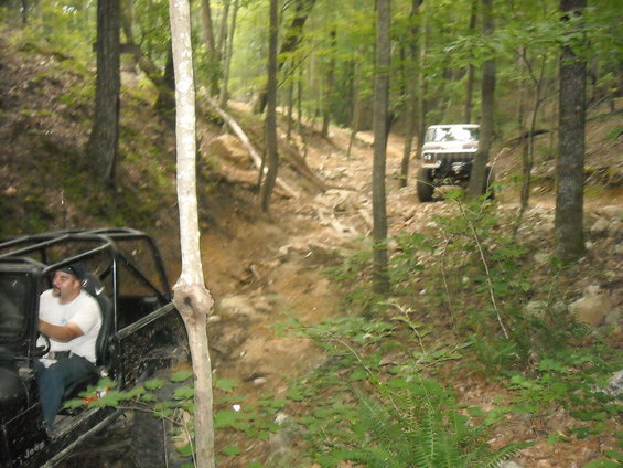 Two off-road vehicles on a rocky, wooded trail. The foreground vehicle is black with a roll cage and a driver. The background vehicle is white