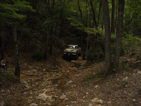 Tan off-road truck with camouflage hood driving on a rocky, wooded trail
