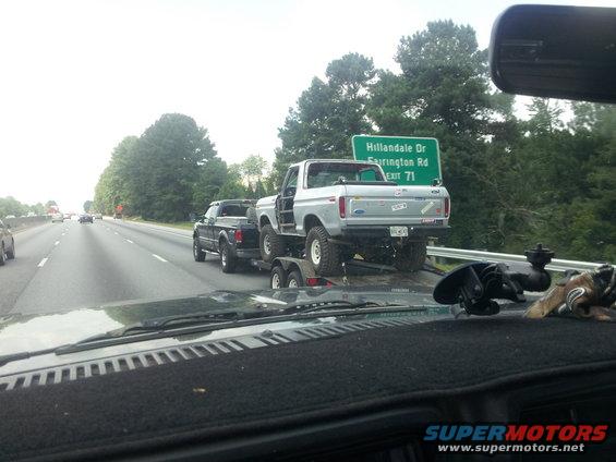 Silver Ford Bronco being towed on a trailer by a black pickup truck on the highway
