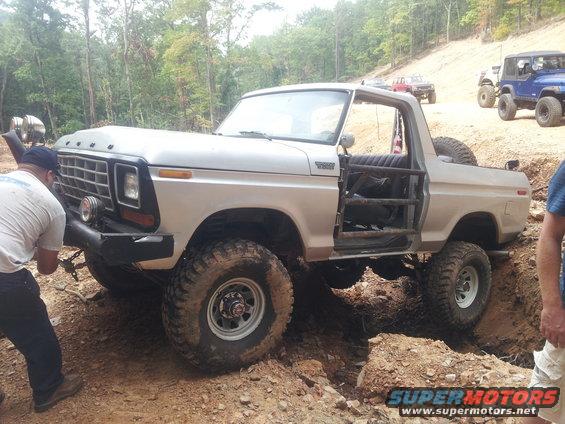 Modified 1978 Ford Bronco with roll cage and off-road tires navigating a dirt obstacle, other off-road vehicles visible on a hill
