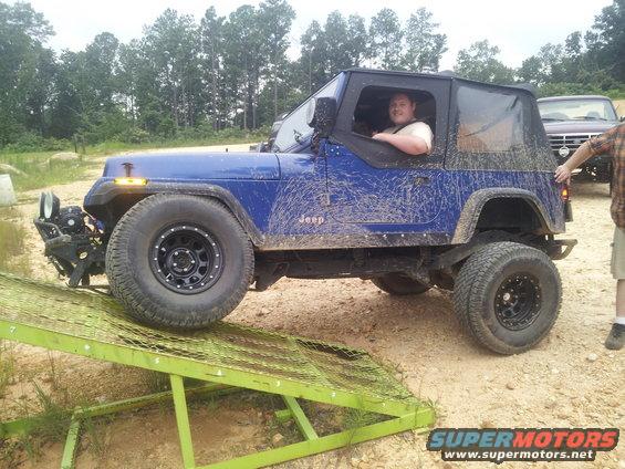 Blue Jeep Wrangler with mud splatters on a green metal ramp, driver visible
