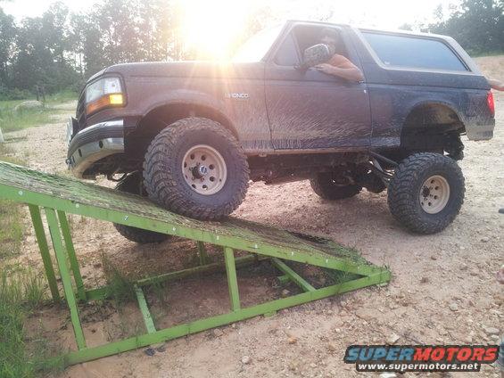 Muddy 1990s Ford Bronco with lifted suspension driving up a green metal ramp
