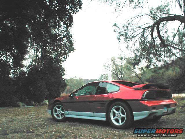 Red 1986 Pontiac Fiero parked on gravel, rear quarter view