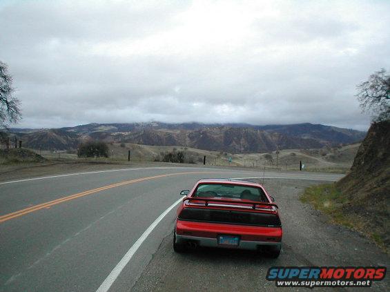 Red 1986 Pontiac Fiero parked on the side of a road with hills in the background