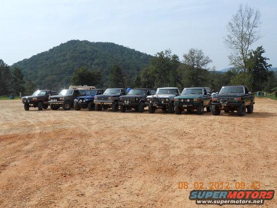 Line of modified off-road trucks and SUVs, mostly from the 1980s, parked on a dirt lot with a hill in the background
