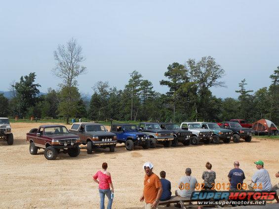 Several lifted trucks and SUVs parked on dirt lot with trees in background. One maroon truck in foreground