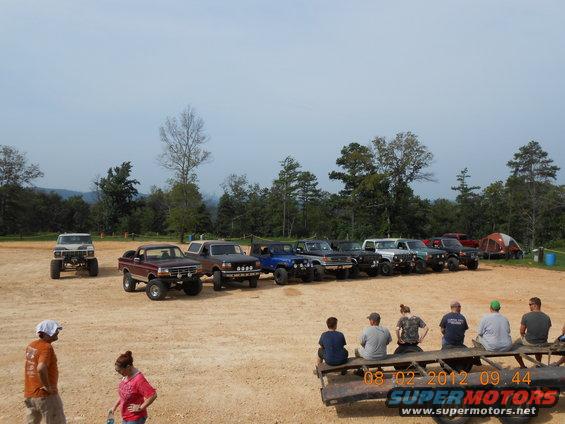 Several off-road trucks and SUVs are parked in a dirt lot with trees and hills in the background. Some people are sitting on a trailer