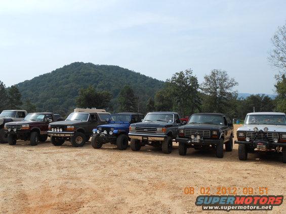 Several lifted Ford trucks and SUVs parked on dirt, with a wooded hill in the background