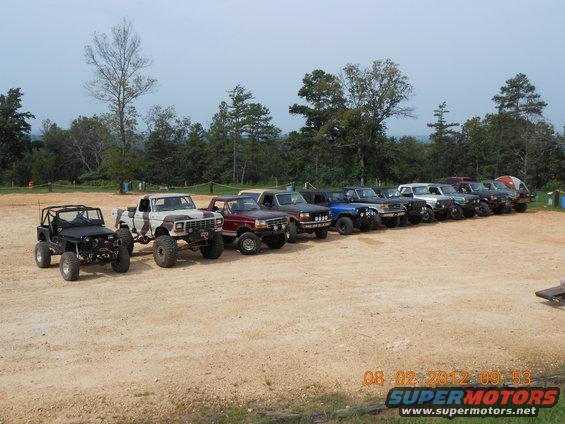 Line of modified off-road trucks and SUVs parked on a dirt lot, with trees and hills in the background