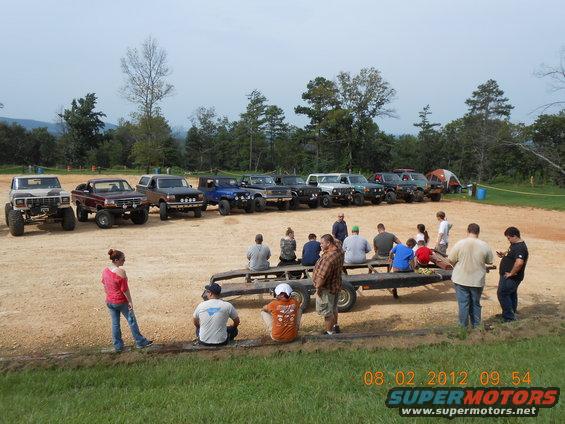 Several off-road trucks and SUVs parked in a dirt lot with people sitting on a trailer and standing around. Date stamp reads 08 02 2012