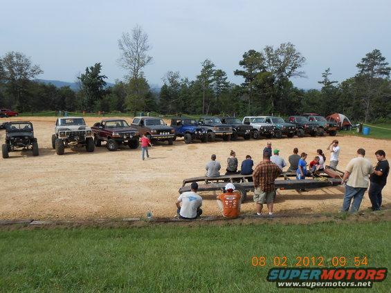 Several off-road trucks and SUVs parked in a dirt lot with people gathered around