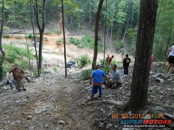 People gathered on a rocky trail with a Jeep driving up a dirt path in the background