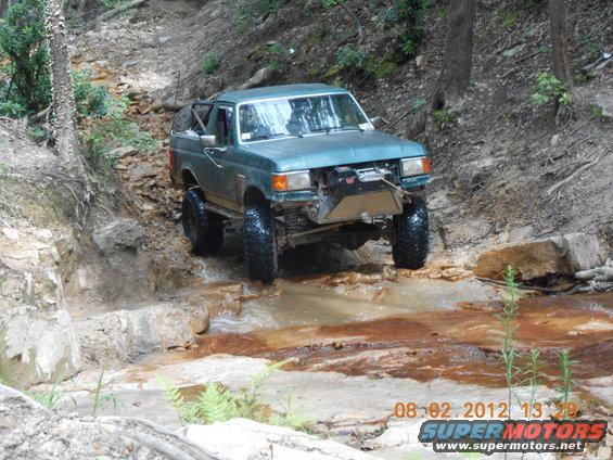 Modified green SUV with large tires navigating a rocky stream, winch on front bumper