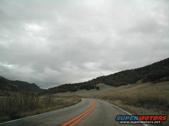 Winding asphalt road with double orange lines curves through a valley under a cloudy sky