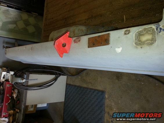 Close-up of a light-colored truck body panel with a red magnetic welding holder attached. There's a small rectangular metal plate and some welded patches visible