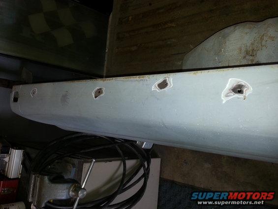 close-up of a light gray vehicle body panel with several holes drilled into it, clamped in a vise