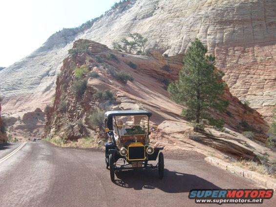 Black 1912 Ford Model T driving on a paved road with a large rock formation and trees