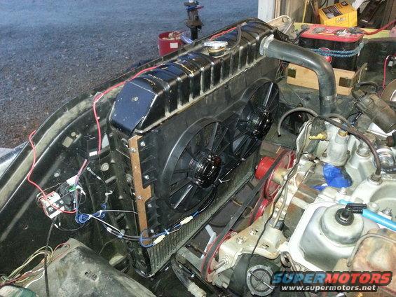 Engine bay showing a radiator with dual electric fans installed, connected by wiring. Parts of the engine block and belts are visible