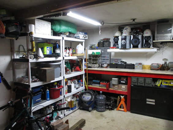 Garage workshop with shelves filled with supplies, tools, and storage bins. A workbench with equipment and a black filing cabinet are visible