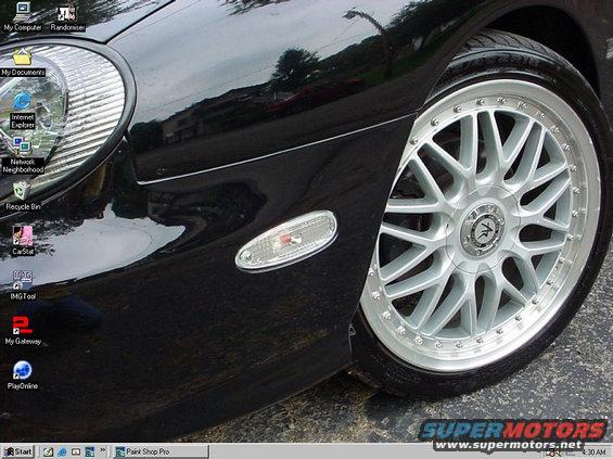 Close-up of a black car's front fender and wheel with aftermarket rims. Computer desktop icons are visible on the screen reflection