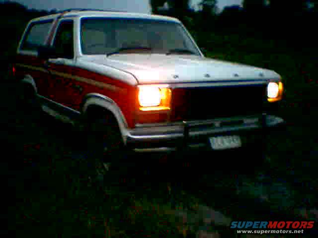 White and red Ford Bronco with its headlights on, parked in a grassy area at dusk
