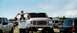 1979 Ford Bronco - A man carries a child on his shoulders in front of a large truck, with other vehicles visible in the background.