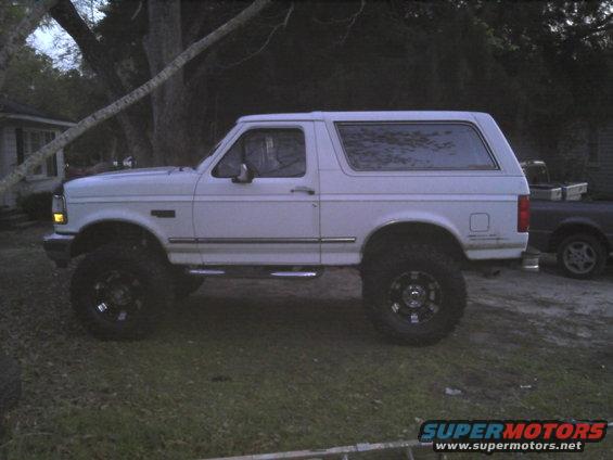 Lifted white SUV with aftermarket wheels parked on grass. Another dark truck is visible behind it