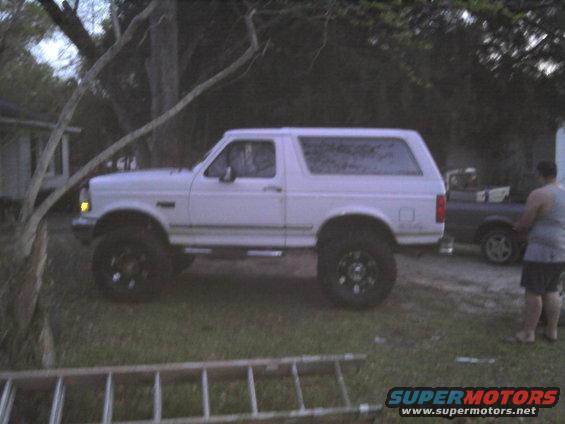 White Ford Bronco, lifted, with black wheels, parked on grass. A person stands near a dark colored car in the background