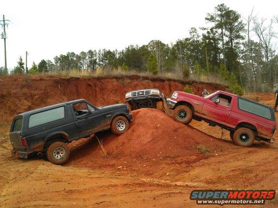 Three Ford Broncos climbing a dirt hill. One is black, one is red, and one is white