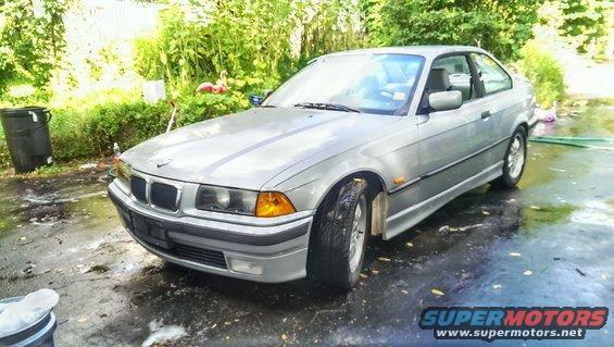 Silver 1997 BMW 3 Series coupe parked on wet asphalt, with trees and bushes in the background